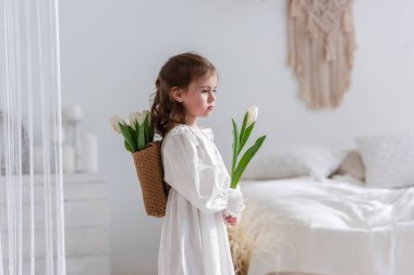 Close-up portrait of a little girl in a white dress, nightgown. Toddler embraces a bouquet of fresh, delicate white tulips. Gift for the holiday, the concept of purity, spring time. Copy space