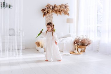 Little girl in white dress, nightgown dancing with back, faceless, on shoulders is hand-made backpack made of raffia with tulips. Light airy, spring interior in rustic style. Pampas grass on the wall