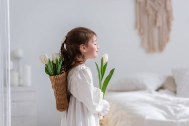 Close-up portrait of a little girl in a white dress, nightgown. Toddler embraces a bouquet of fresh, delicate white tulips. Gift for the holiday, the concept of purity, spring time. Copy space