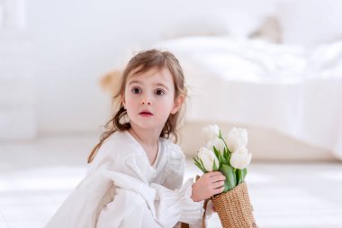 Close-up portrait of a little girl in a white dress, nightgown. Toddler embraces a bouquet of fresh, delicate white tulips. Gift for the holiday, the concept of purity, spring time. Copy space