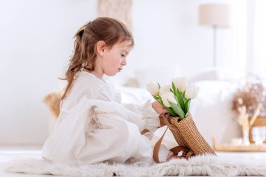 Close-up portrait of a little girl in a white dress, nightgown. Toddler embraces a bouquet of fresh, delicate white tulips. Gift for the holiday, the concept of purity, spring time. Copy space