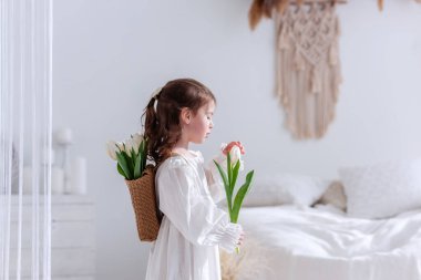 Close-up portrait of a little girl in a white dress, nightgown. Toddler embraces a bouquet of fresh, delicate white tulips. Gift for the holiday, the concept of purity, spring time. Copy space