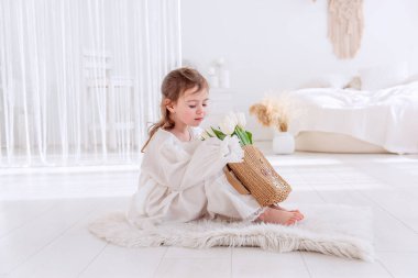 Beyaz Boho minimalist iç mekanında küçük bir kız yerde oturuyor. Bebek elbiseli, gecelikli, elinde bir buket bahar lalesi tutan, sarılan, çiçek kokan. Airy, ışık dolu bir yer. Pampas çimen dekoru