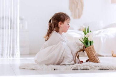 Beyaz Boho minimalist iç mekanında küçük bir kız yerde oturuyor. Bebek elbiseli, gecelikli, elinde bir buket bahar lalesi tutan, sarılan, çiçek kokan. Airy, ışık dolu bir yer. Pampas çimen dekoru