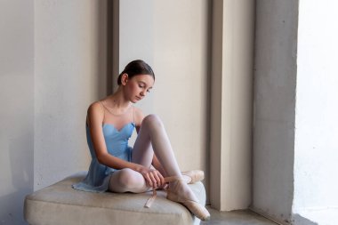 Genç, dingin balerin kız, puantiyeli ayakkabılar takarak koltuğa oturur. Güneş ışığı dikey pencerelerden akar, yumuşak gölgeler bırakır, dans stüdyosunun sessiz atmosferini güçlendirir.