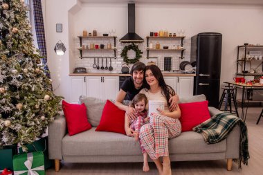 Noel için ağaç ve hediyelerle süslenmiş rahat, şenlikli bir oturma odasında küçük kızıyla gülümseyen ebeveynler. Modern İskandinav iç mimarisi. Küçük kız oynaşıyor, yüzünü buruşturuyor.