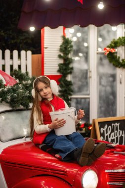 Kulaklık takan genç bir kız, elinde çelenkle klasik bir arabada oturuyor ve hediyesini açıyor. Noel süslemeleri ve ışıklar dışarıda sıcak bir tatil ortamı yaratıyor. Arka bahçe.
