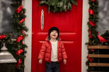 Kırmızı ceketli neşeli çocuk, çelenk ve çelenklerle süslenmiş, şenlikli kırmızı kapının önünde duruyor. Noel ağacı ve ışıklar arka bahçede rahat bir tatil ortamı yaratıyor.