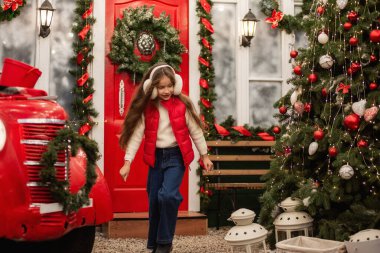 Kulaklık takan ve kırmızı yelek giyen genç bir kız klasik bir arabanın yanından mutlu bir şekilde geçiyor ve bir çelenk ve süs de dahil olmak üzere Noel süslemeleri kutluyor. Tatil sahnesi sıcaklık ve neşeyle parlıyor..