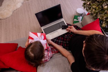 İnternetten alışveriş yapmak için dizüstü bilgisayar ve kredi kartı kullanan bir baba ve kızın elinde tatil dekoruyla sarılı bir hediye tutarken görülüyor..