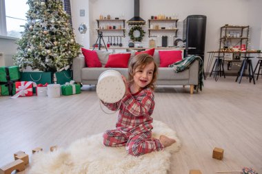 Ekose pijamalı küçük bir çocuk süslü oturma odasında pofuduk beyaz bir halının üzerinde oturuyor, elinde gülümseyerek yuvarlak bir hediye kutusu tutuyor. Noel ağacı süslerle, ışıklarla, hediye kutularıyla, rahat mobilyalarla süslenmiş..