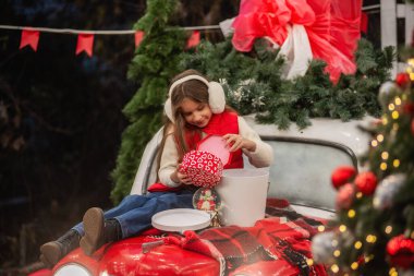 Kulaklık takan genç bir kız, elinde çelenkle klasik bir arabada oturuyor ve hediyesini açıyor. Noel süslemeleri ve ışıklar dışarıda sıcak bir tatil ortamı yaratıyor. Arka bahçe.