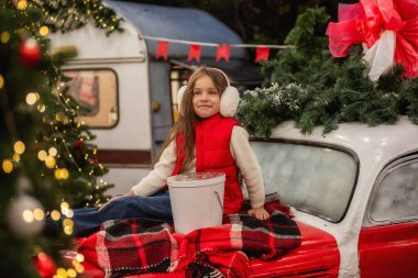 Kulaklık takan genç bir kız, elinde çelenkle klasik bir arabada oturuyor ve hediyesini açıyor. Noel süslemeleri ve ışıklar dışarıda sıcak bir tatil ortamı yaratıyor. Arka bahçe.