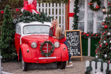 Süslü kırmızı bir arabaya yaslanmış gülümseyen bir kadın. Mutlu Yıllar tabelası olan, etrafı şenlikli bir dekorla çevrili. Kutlama havasını şenlikli yeşillik ve tatil dekoru belirler.