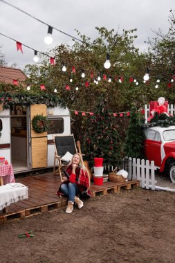 Woman in cozy plaid blanket sits outside a holiday-decorated camper. Christmas tree, guitar, and rustic holiday decor create a warm and inviting festive atmosphere. clipart