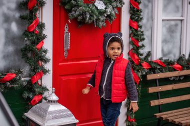 Sıcak bir ayı kapüşonlu ve kırmızı yelekli neşeli çocuk Noel çelenkleri ve çelenklerle süslenmiş neşeli kırmızı kapıyı çalar ve Noel mevsiminin büyüsünü yakalar..
