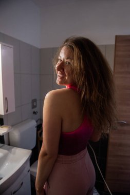 Pembe spor bluzlu neşeli kadın saçlarını loş bir banyoda kuruturken arkasına bakıyor. Kendine gülerek sıcak ve rahat bir atmosfer yaratıyor. Rock yıldızı