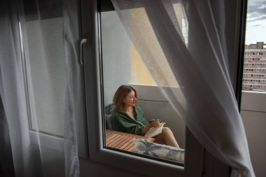 Yeşil ekose elbiseli kızıl saçlı kadın balkonuna oturmuş, sessiz ve düşünceli bir anın tadını çıkarırken bir deftere bir şeyler yazıyor. Yumuşak perdeler, huzurlu açık hava sahnesini çerçeveler..