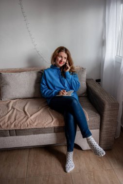 Mavi kapüşonlu ve benekli çoraplı genç bir kadın rahat bir koltukta oturur, elinde bir defter tutarken telefonla konuşur. Rahatlamış ve sohbete dalmış görünüyor..