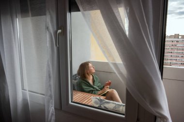 Yeşil ekose elbiseli genç bir kadın küçük bir balkonda huzurlu bir anın tadını çıkarıyor, elinde bir kitapla dışarıya bakıyor. Yumuşak doğal ışık perdelerin arasından süzülür.