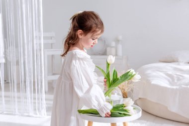 Gecelikli, parlak, güneşli bir odada beyaz laleleri düzenleyen, çiçek bakımının huzurlu anının tadını çıkaran ve bahar tazeliğinin güzelliğini kucaklayan sevimli bir çocuk. Boho iç mimarisi