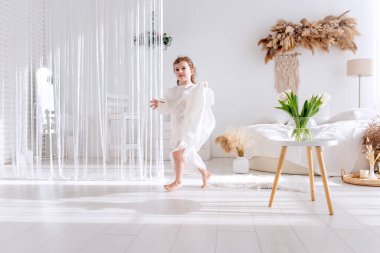 Beyaz bir elbise giyen, uzun kahverengi saçlı tatlı küçük kız bohem dekorasyonlu, otlu ve beyaz laleli beyaz yatak odasında yalınayak koşuyor.