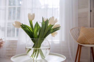 On round wooden table stands glass vase with a bouquet of white tulips. Airy, light interior in bright colors. Freshness, purity of natural colors. Spring time. Flowers for a holiday, mood. Copy space