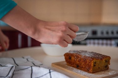 Şekerci, sıcak bir mutfakta, ahşap bir tahtanın üzerinde yeni pişmiş bir somun keki üzerine şeker serpiyor. Tatlı ikram servis edilmeye hazır. Ev yapımı yemek.