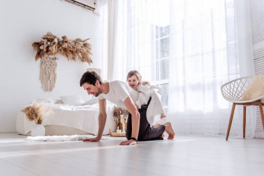 Mutlu baba, küçük kızı sırtında şınav çekmekten zevk alıyor, neşeli anları paylaşıyor ve doğal ışıkla dolu, parlak, rahat bir yatak odasında birlikte egzersiz yapıyor.