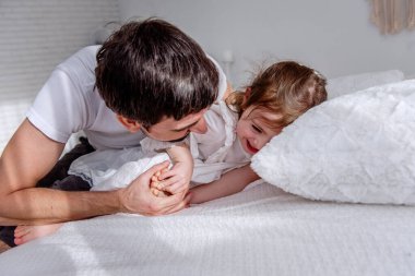 Baba ve kızı sıcak bir yatakta neşeli bir anı paylaşıyorlar, aile sevgisinin ve bağlantının özünü yakalıyorlar kahkaha ve şefkatle dolu samimi bir sahnede.