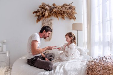 Baba ve kızı bir yatakta oturuyor, taş kağıt makas oynuyorlar, sıcacık bir bohem yatak odasında, kurumuş çiçekler ve makrome ile güzel vakit geçiriyorlar.