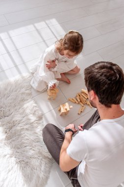 Babası ve küçük kızı beyaz ahşap bir zeminde, yumuşak bir halının yanında oturuyorlar, tahta bloklarla oynuyorlar, küçük yapılar inşa ediyorlar ve birlikte eğleniyorlar.