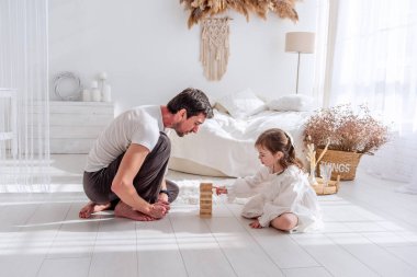 Baba ve kızı beyaz bir yatak odasında yerde oturmuş, ahşap bir kule oyununa konsantre olup, birlikte güzel vakit geçiriyorlar.