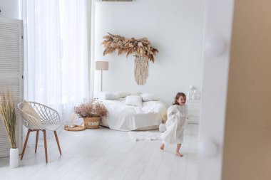 Beyaz elbiseli, uzun kahverengi saçlı neşeli küçük kız bohem dekorasyonlu, rahat beyaz bir yatak odasında yalınayak koşuyor, saklambaç oynuyor.
