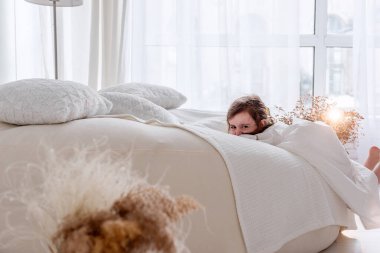 Mutlu küçük kız parlak bir yatak odasında saklambaç oynuyor, rahat bir yatağın arkasına saklanıyor, evde neşeli bir anın tadını çıkarıyor.