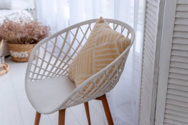 Sarı yastıklı, rahat, modern, beyaz bir sandalye. Ahşap bacaklı, beyaz perdeli, parlak bir odada rahat bir atmosfer yaratıyor.