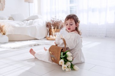 Beyaz elbiseli şirin küçük kız, beyaz bir yatak odasında yerde oturuyor, elinde beyaz lalelerle dolu bir torba tutuyor.