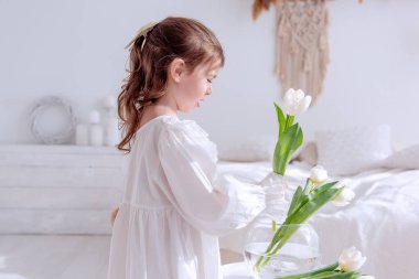 Gecelikli, parlak, güneşli bir odada beyaz laleleri düzenleyen, çiçek bakımının huzurlu anının tadını çıkaran ve bahar tazeliğinin güzelliğini kucaklayan sevimli bir çocuk. Boho iç mimarisi
