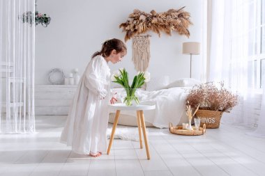 Beyaz elbiseli genç kız, rahat bir yatağı ve doğal dekorasyonları olan, parlak, bohem tarzı bir yatak odasında, küçük bir masanın üzerine yerleştirilmiş taze beyaz lalelerin kokusunu almak için eğiliyor.