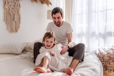 Mutlu baba, rahat bir yatakta rahat bir şekilde otururken neşeli kızını gıdıklıyor. Sıcak, güneşli bir yatak odasında. Yürek ısıtan bir aile bağı ve eğlenceli bir etkileşim anı yaratıyor.