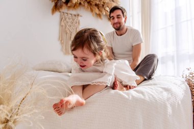 Mutlu baba, rahat bir yatakta rahat bir şekilde otururken neşeli kızını gıdıklıyor. Sıcak, güneşli bir yatak odasında. Yürek ısıtan bir aile bağı ve eğlenceli bir etkileşim anı yaratıyor.
