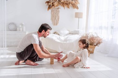 Baba ve kızı beyaz bir yatak odasında yerde oturmuş, ahşap bir kule oyununa konsantre olup, birlikte güzel vakit geçiriyorlar.