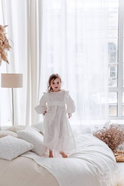 Uzun kahverengi saçlı neşeli küçük kız büyük, rahat bir yatakta beyaz bir yatak odasında uzun beyaz bir elbise giyerek neşeli bir anın tadını çıkarıyor.