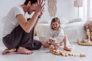 Babası ve küçük kızı yerde oturup neşe içinde ahşap bloklarla oynuyorlar. Kahkahalarını paylaşıyorlar ve samimi yatak odalarında güzel anılar yaratıyorlar.