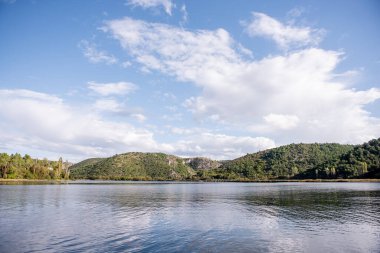 Visovac gölü, güneşli bir günde Hırvatistan 'ın Krka ulusal parkında güzel bir manzara olan yemyeşil bitki örtüsü ve tepelerle çevrili sakin sularıyla bulutlu gökyüzünü yansıtıyor.