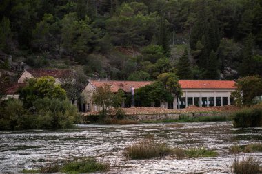 Hırvatistan 'ın Krka milli parkı içinde yemyeşil bitki örtüsü ve ormanlı tepelerle çevrili Krka nehrinin yanında duran eski hidroelektrik santrali binaları
