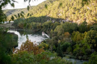 Hırvatistan 'ın Krka ulusal parkında, yemyeşil ağaçlar ve yumuşak su akıntılarıyla çevrili sakin Krka nehrinde dinlenen bir turist teknesinin yer aldığı sakin bir manzara