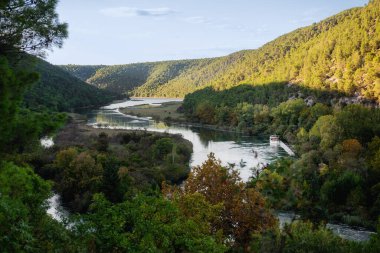 Hırvatistan 'ın Krka ulusal parkındaki sakin manzarada, yemyeşil ve sakin sularla çevrili dolambaçlı Krka nehrinde gezinen bir turist botu yer alıyor.