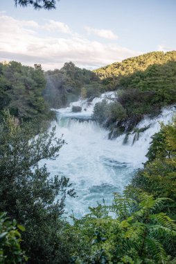 Hırvatistan 'daki Krka ulusal parkının çağlayan şelalelerini çevreleyen yemyeşil bitki örtüsü bulutlu bir gökyüzü altında nefes kesici bir doğal manzara yaratıyor.