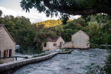 Hırvatistan 'ın Krka milli parkındaki, yemyeşil ağaçlar ve tepelerle çevrili eski taş su değirmenlerinin etrafında akan sular, tarihi sanayi ve doğal güzelliğin sakin ve resimsel bir manzarasını oluşturuyor.